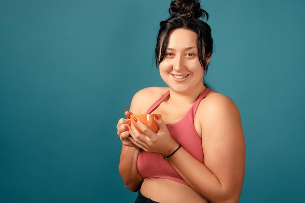 Feliz mujer positiva de talla grande. Concepto positivo de cuerpo feliz. Me encanta mi cuerpo. Atractiva mujer con sobrepeso posando en la cámara en el estudio con una taza de té en las manos. La chica lleva un sostén de coral.