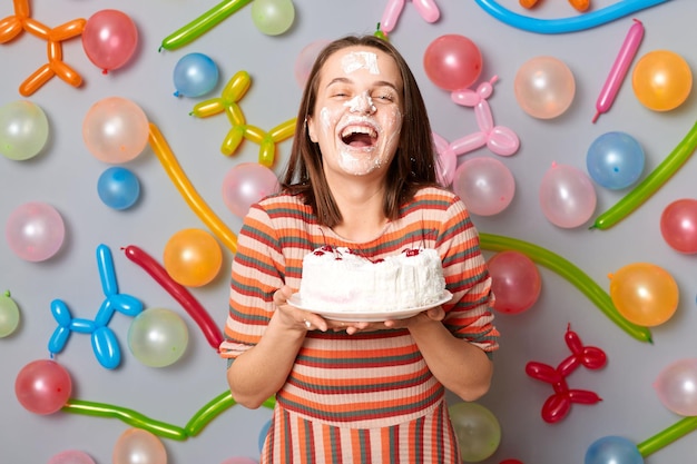 Feliz mujer riendo alegre con vestido a rayas de pie contra la pared gris decorada con globos de colores celebrando el cumpleaños con pastel con crema en la cara