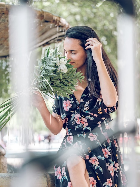 Feliz mujer romántica sentada al borde de la fuente y disfrutando del aroma del ramo de flores presentado en el día de verano