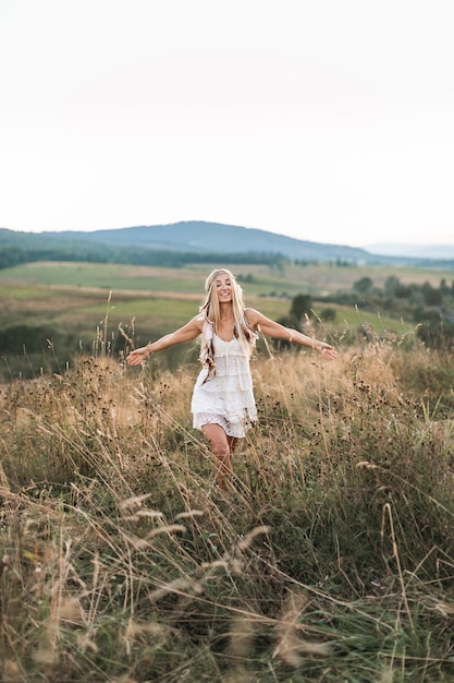 Feliz mujer rubia atractiva boho en vestido blanco y accesorios de plumas en el cabello en el campo de verano al aire libre