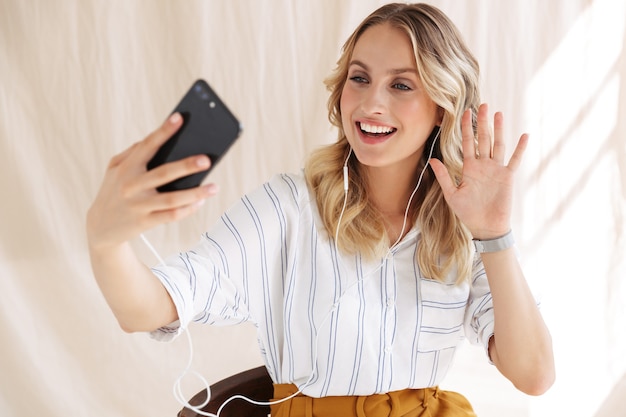 Feliz mujer rubia con auriculares saludando al smartphone en la mano