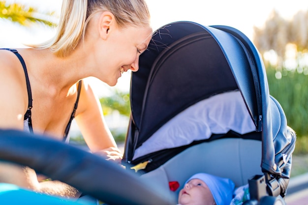 Feliz mujer rubia besándose y sintiéndose adorada por su paseo por un niño recién nacido en el parque tropical de la playa al aire libre