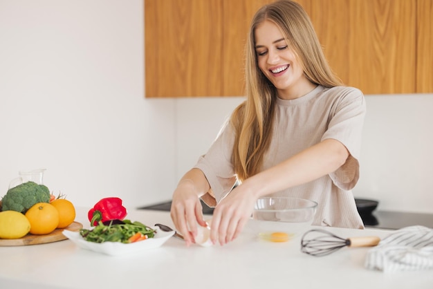 Feliz mujer rubia bonita en pijama está preparando el desayuno de huevos y verduras frescas en la cocina en casa