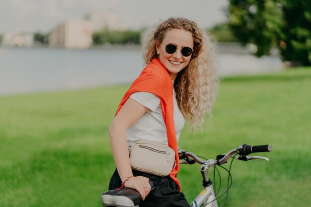 Feliz mujer rubia de cabello rizado disfruta de un paseo en bicicleta tiene su propio transporte lleva un estilo de vida saludable viaja en bicicleta alrededor de vistas panorámicas de la naturaleza vestida con ropa informal tiene vacaciones activas