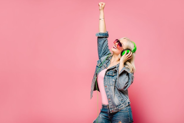 Feliz mujer rubia con estilo en gafas rosas con grandes auriculares escuchando música y bailando sobre fondo de color