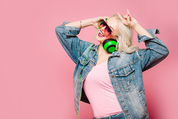 Feliz mujer rubia con estilo en gafas rosas con grandes auriculares escuchando música y bailando sobre fondo de color