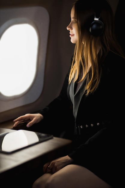 Feliz mujer rubia joven caucásica en elegante vestido negro relajante durante el vuelo en jet privado, escuchando música con auriculares inalámbricos con los ojos cerrados.