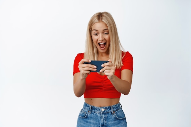 Feliz mujer rubia jugando videojuegos en el teléfono, mirando en el teléfono inteligente con asombrada expresión de la cara sonriente en blanco