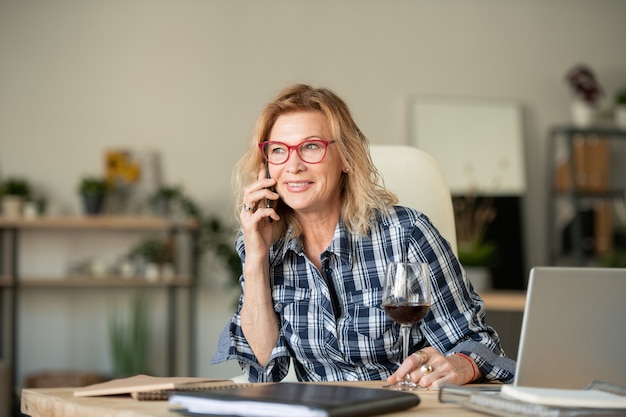 Feliz mujer rubia madura con smartphone y copa de vino tinto sentado en el escritorio frente a la computadora portátil mientras habla por teléfono en casa