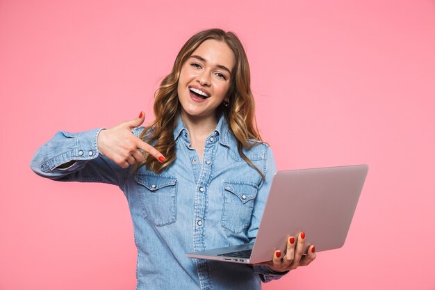 Feliz mujer rubia vestida con camisa de mezclilla sosteniendo la computadora portátil y apuntando mientras mira al frente sobre la pared rosa