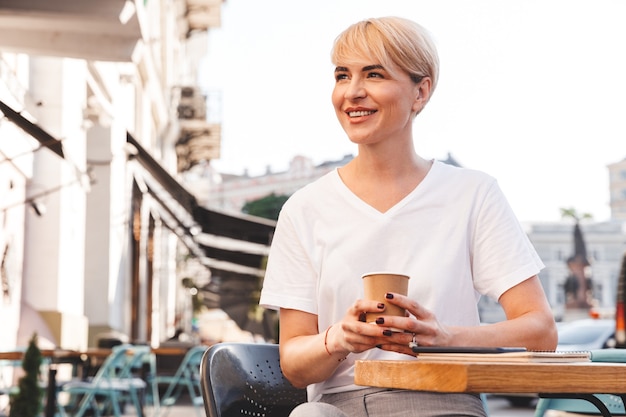 Feliz mujer rubia vestida con camiseta blanca sentada en la cafetería de la calle en verano y bebiendo café de la taza de papel