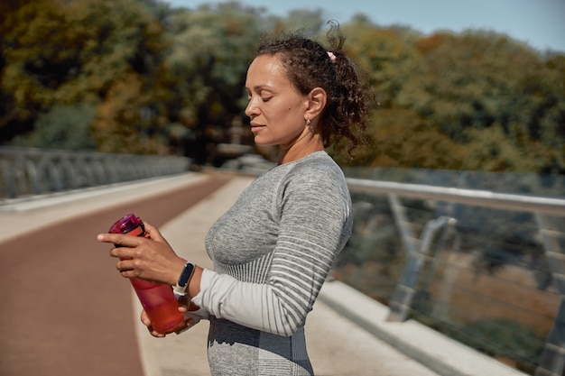 Feliz mujer sana y deportiva está bebiendo agua después de los entrenamientos de entrenamiento en un día soleado