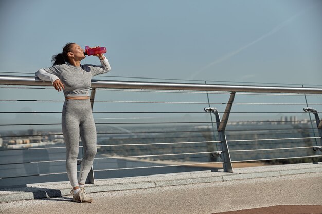 Feliz mujer sana y deportiva está bebiendo agua después de los entrenamientos de entrenamiento en un día soleado