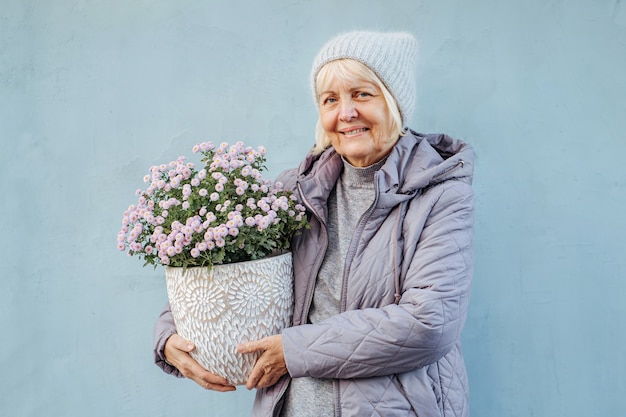 feliz mujer senior en ropa de abrigo sonriendo y llevando flores en macetas el día de la primavera.