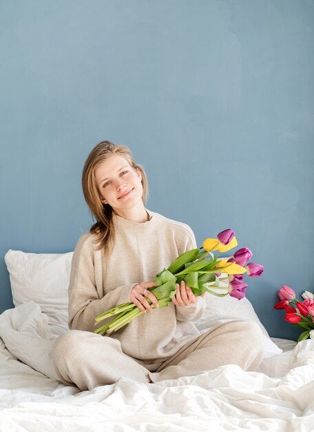 Feliz mujer sentada en la cama en pijama con ramo de flores de tulipán, fondo de pared azul