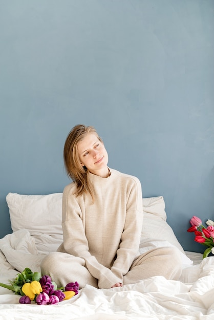 Feliz mujer sentada en la cama en pijama con ramo de flores de tulipán, fondo de pared azul