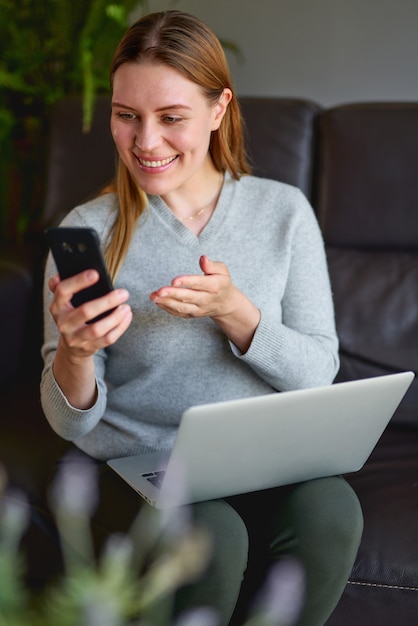 Feliz mujer sentada en el sofá con el portátil y hablando por teléfono en casa. Estudiante universitario que estudia en la computadora portátil y que usa el teléfono.