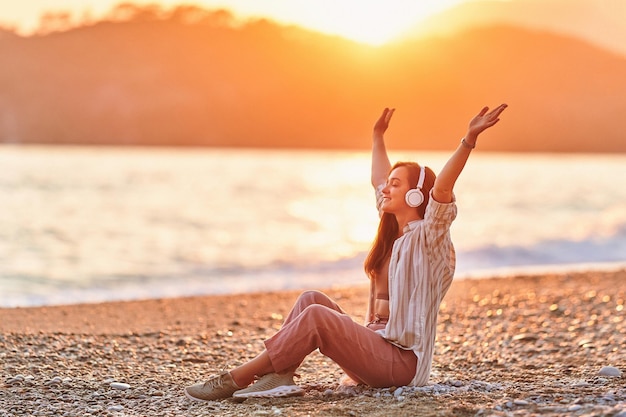 Feliz mujer sola con los brazos abiertos y los ojos cerrados disfruta de escuchar música relajante en la orilla del mar al atardecer