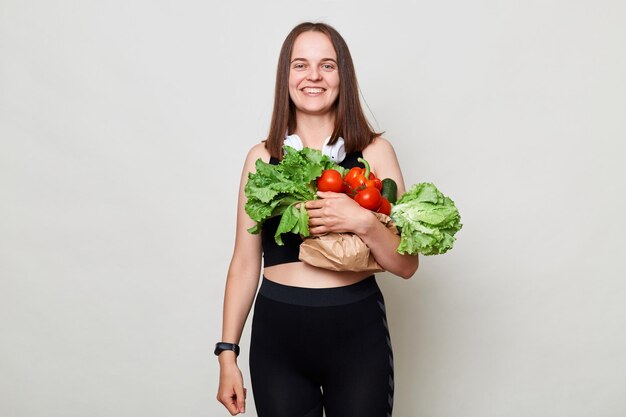 Feliz mujer sonriente y alegre con cabello oscuro lleva verduras compradas en el mercado aislada sobre un fondo gris disfrutando de una alimentación saludable mirando a la cámara con cara satisfecha