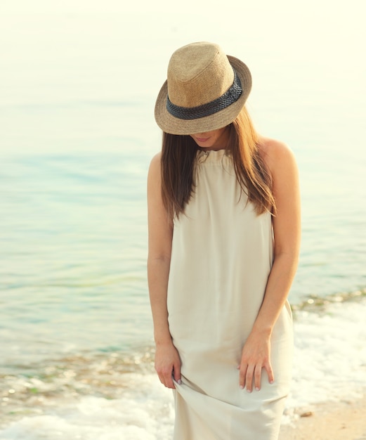 Feliz mujer sonriente caminando por una playa de mar vestida con vestido blanco y sombrero que cubre la cara, relajarse y disfrutar del aire fresco.