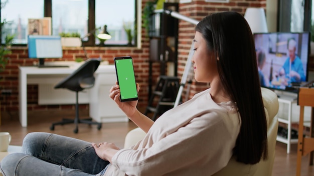 Feliz mujer sonriente de corazón que tiene un teléfono inteligente con pantalla verde mientras se sienta en casa en el sofá. Una persona alegre sentada en un sofá con un teléfono móvil con un fondo de clave de croma de maqueta.