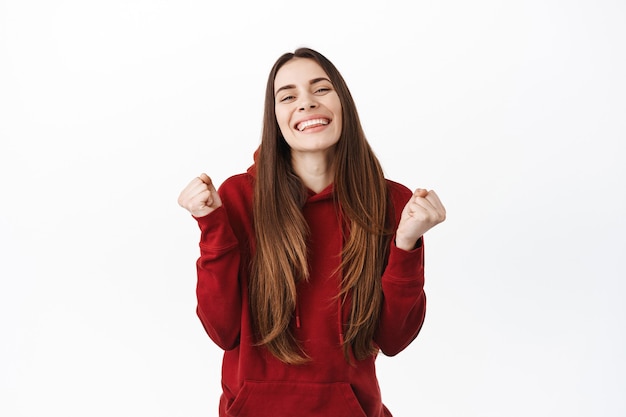 Feliz mujer sonriente ganando y celebrando, lucir encantada con bombas de puño, decir que sí, lograr la meta, sentir el dulce sabor del éxito, convertirse en campeona, de pie sobre una pared blanca