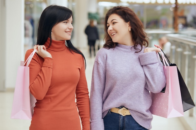 Feliz mujer sonriente mirando el uno al otro mientras hace compras en el centro comercial, posando con bolsas