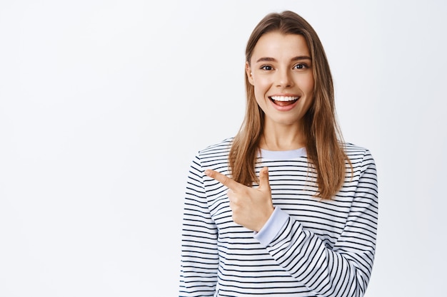 Feliz mujer sonriente con pelo corto rubio, apuntando con el dedo a la izquierda en el espacio de la copia, mostrando el camino o la dirección del anuncio, de pie sobre una pared blanca