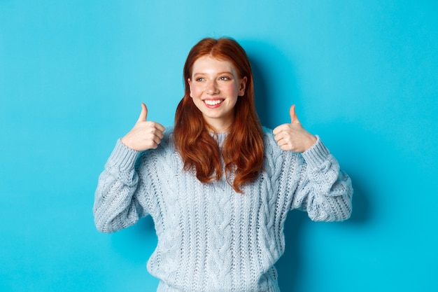 Feliz mujer sonriente con el pelo rojo, mostrando los pulgares hacia arriba y mirando a la izquierda satisfecho, alabando la buena elección, de acuerdo y aprobar, de pie sobre fondo azul.