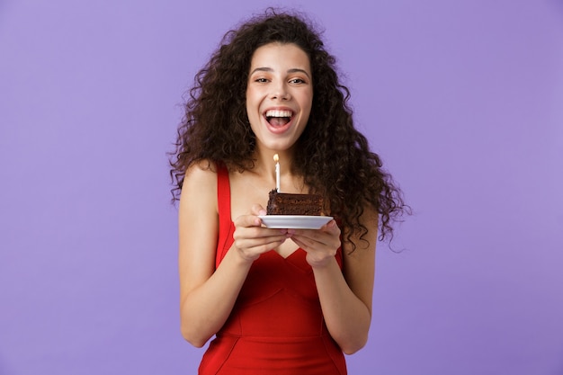 Feliz mujer vistiendo un vestido rojo sosteniendo un trozo de tarta de cumpleaños con vela, aislado sobre pared violeta