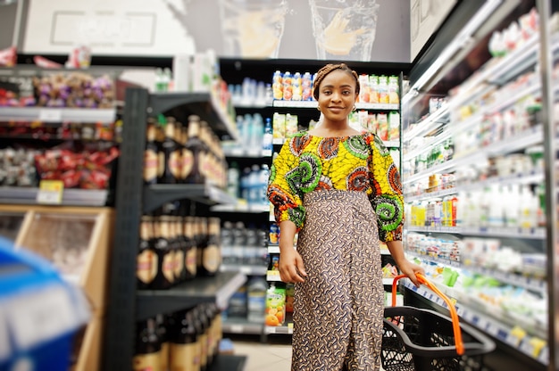 Feliz mulher africana em roupas tradicionais e véu, olhando o produto na mercearia, compras no supermercado. Cliente afro das mulheres negras com cesta que compra comida no mercado.