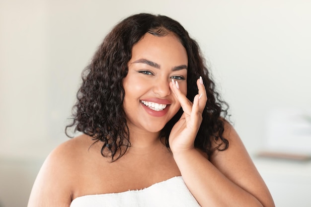 Feliz mulher afro-americana plus size sorrindo para a câmera e tocando o nariz aplicando produtos faciais após o banho