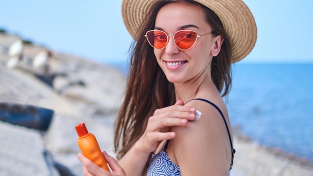 Feliz mulher alegre em traje de banho, chapéu de palha e óculos de sol vermelhos brilhantes aplicar protetor solar no ombro durante o banho de sol e relaxar à beira-mar