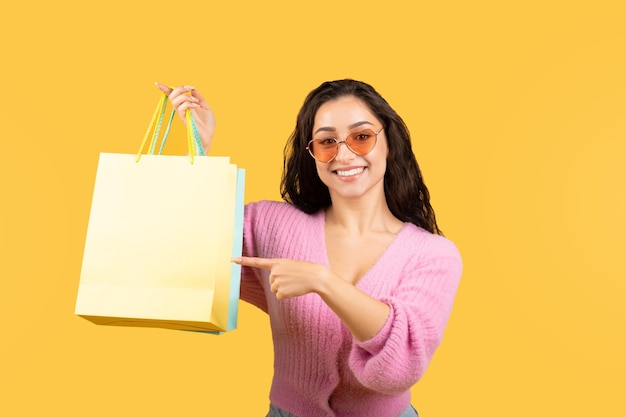 Feliz mulher árabe milenar em óculos de sol e dedo apontando casualmente para pacotes recomenda fazer compras