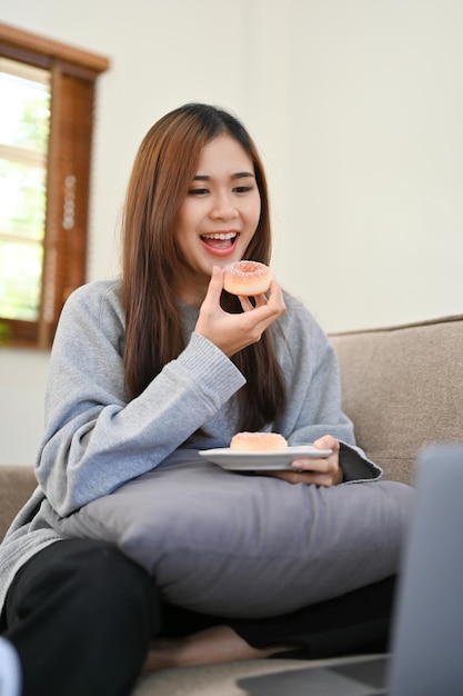 Feliz mulher asiática comendo rosquinha na sala de estar enquanto relaxa