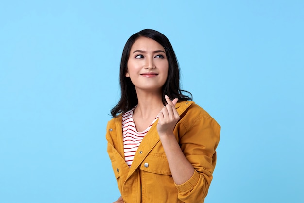 Feliz mulher asiática em camisa amarela e mini coração de gesto de mão sobre o espaço azul.