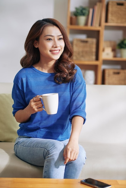 Feliz mulher asiática jovem e atraente com cabelo encaracolado, sentada com uma caneca no sofá e dando uma entrevista ou conversando com alguém