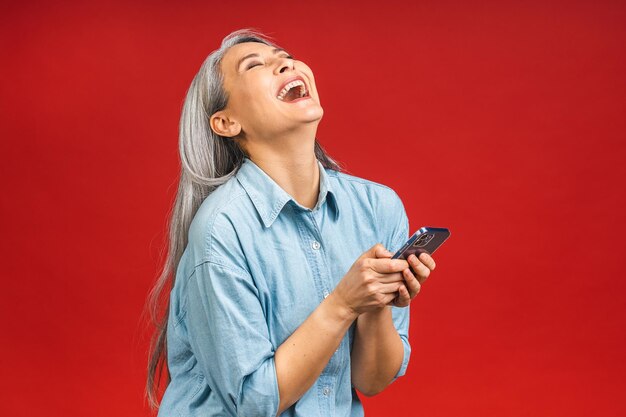 Feliz mulher asiática sênior madura segurando smartphone usando aplicativos móveis online sorrindo velha senhora de meia idade mandando mensagem sms conversando no telefone isolado sobre fundo vermelho