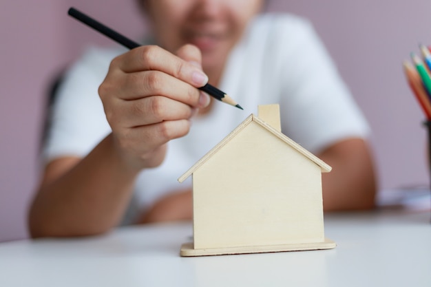 Feliz mulher asiática usando lápis para desenhar com casa de madeira cofrinho metáfora design e decoração a casa selecione foco profundidade de campo rasa