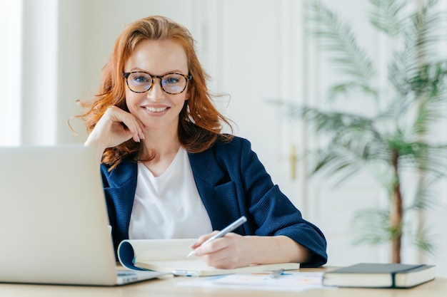 Feliz mulher bem sucedida escreve lista de verificação no bloco de notas trabalha em ideias criativas para pesquisas de projeto de inicialização informações necessárias em poses de computador portátil no espaço de coworking trabalha freelance