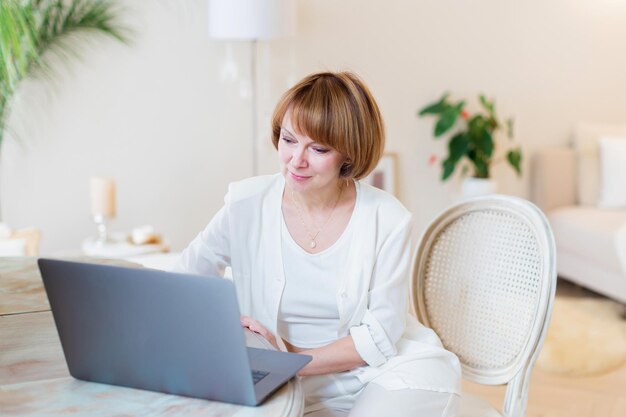 Feliz mulher bonita casual trabalhando em um laptop sentado na cadeira na elegante sala de estar branca