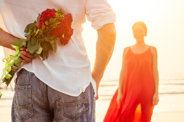 Feliz mulher bonita de vestido vermelho recebendo um presente de um homem por mar por do solsurpresa esposa do marido buquê de rosas dia da mulher 8 de março e dia dos namorados Noivado amor à primeira vista romântico