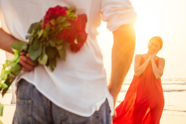 Feliz mulher bonita de vestido vermelho recebendo um presente de um homem por mar por do solsurpresa esposa do marido buquê de rosas dia da mulher 8 de março e dia dos namorados Noivado amor à primeira vista romântico