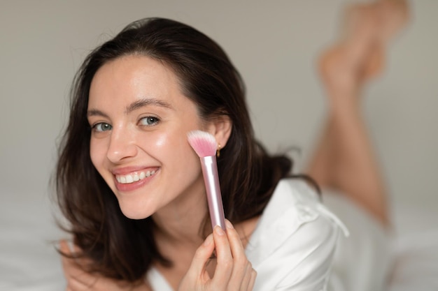 Feliz mulher caucasiana do milênio aplicando pó com pincel no rosto está na cama