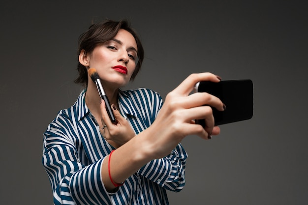 Feliz mulher caucasiana, maquiagem com pincéis e tirar uma selfie na parede cinza