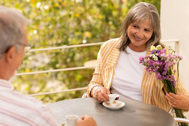 Feliz mulher caucasiana sênior com buquê, olha para o homem bebe café, senta-se à mesa no café