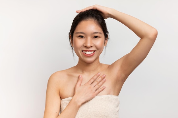 Feliz mulher coreana sensual embrulhada em toalha branca, levantando a mão mostrando-a limpa e sem pelos