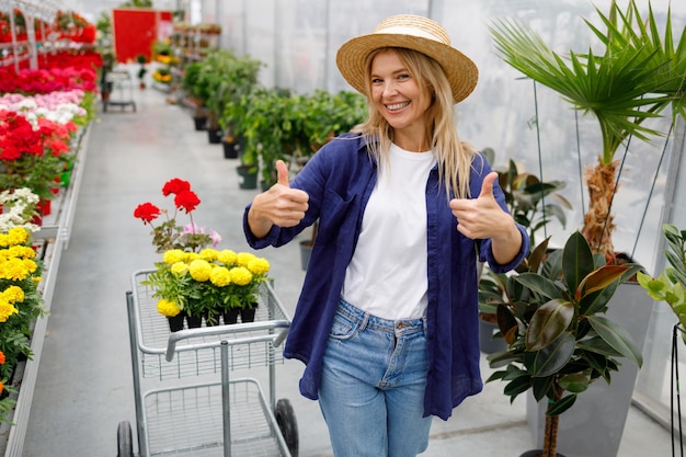 Feliz mulher de chapéu com carrinho de compras mostrando os polegares depois de comprar flores