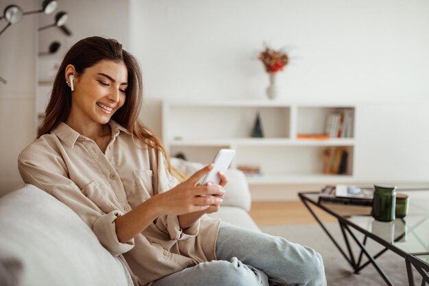 Feliz mulher de raça mista do milênio em fones de ouvido sem fio, olha para o telefone, lê a mensagem conversando