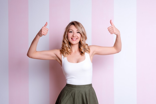 Feliz mulher emocional posando e aparecendo os polegares no fundo rosa. Jovem mulher loira desistindo polegares, aprovando fazendo gesto positivo com a mão para o sucesso. Olhando para a câmera, gesto de vencedor